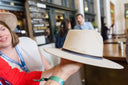 Person holding a sand suede hat with a blurred background of people and indoor setting