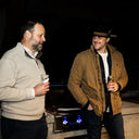 Two men in hats standing next to each other, holding drinks, with a dark background