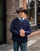 Man wearing a navy sweater and cowboy hat standing against a brick wall.