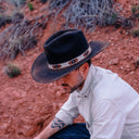 Man wearing a black cowboy hat with a red and white band, sitting on a rocky desert landscape.