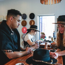 Three people sitting at a table with hats on, in a room with hats displayed on the wall.