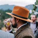 Man in a brown hat and suit drinking from a glass outdoors with mountains in the background