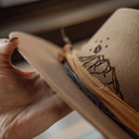 Close-up of a hand holding a brown hat with a branded mountain design.