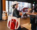 Person holding a tote bag with a red hat and white design in a casual setting