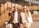 Man and woman in cowboy hats and formal attire walking on a wooden deck at night.