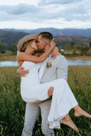 Couple in wedding attire and bridal hat embracing in a field with mountains in the background