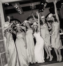 Group of women in dresses and hats posing together in a room with chandeliers.
