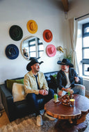 Two men in cowboy hats sitting on a couch in a room with colorful hat decorations on the wall.